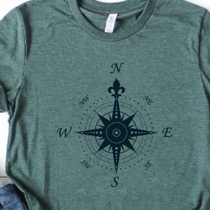 Puede incluir: Una camiseta verde con un dise&ntilde;o de br&uacute;jula azul marino. La br&uacute;jula tiene una flor de lis en la parte superior y las direcciones cardinales est&aacute;n etiquetadas como N, S, E, W, NE, NW, SE y SW.