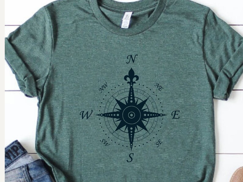 Puede incluir: Una camiseta verde con un dise&ntilde;o de br&uacute;jula azul marino. La br&uacute;jula tiene una flor de lis en la parte superior y las direcciones cardinales est&aacute;n etiquetadas como N, S, E, W, NE, NW, SE y SW.
