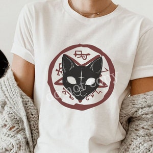 K&ouml;nnte beinhalten: Wei&szlig;es T-Shirt mit einer schwarzen Katzenillustration in einem roten Kreis mit einem Pentagramm und anderen Symbolen. Die Katze hat wei&szlig;e Augen und ein Kreuz auf der Stirn.
