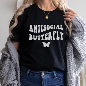 Puede incluir: Una camiseta negra con texto blanco que dice "Antisocial Butterfly" en una fuente retro. Un gr&aacute;fico de mariposa blanca est&aacute; debajo del texto.