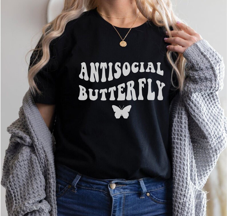 Puede incluir: Una camiseta negra con texto blanco que dice "Antisocial Butterfly" en una fuente retro. Un gr&aacute;fico de mariposa blanca est&aacute; debajo del texto.