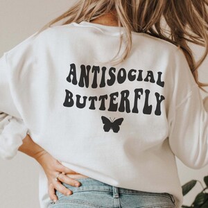 Puede incluir: Sudadera blanca con texto negro que dice "Antisocial Butterfly" en una fuente retro. Un peque&ntilde;o gr&aacute;fico de mariposa negra se encuentra debajo del texto.