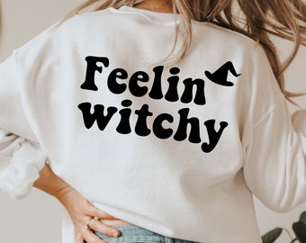 Puede incluir: Sudadera blanca con texto negro que dice "Feelin witchy" con un pequeño sombrero de bruja gráfico sobre la palabra "witchy".