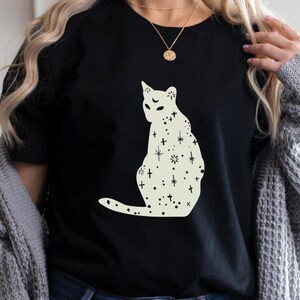 K&ouml;nnte beinhalten: Ein schwarzes T-Shirt mit einer wei&szlig;en Silhouette einer Katze mit Sternen und Kreuzen auf ihrem K&ouml;rper.