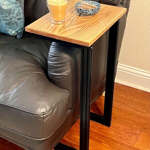 May include: A wooden side table with a light brown tabletop and black metal frame. The table is positioned next to a black leather couch. A glass of coffee and a small bowl are on the table.