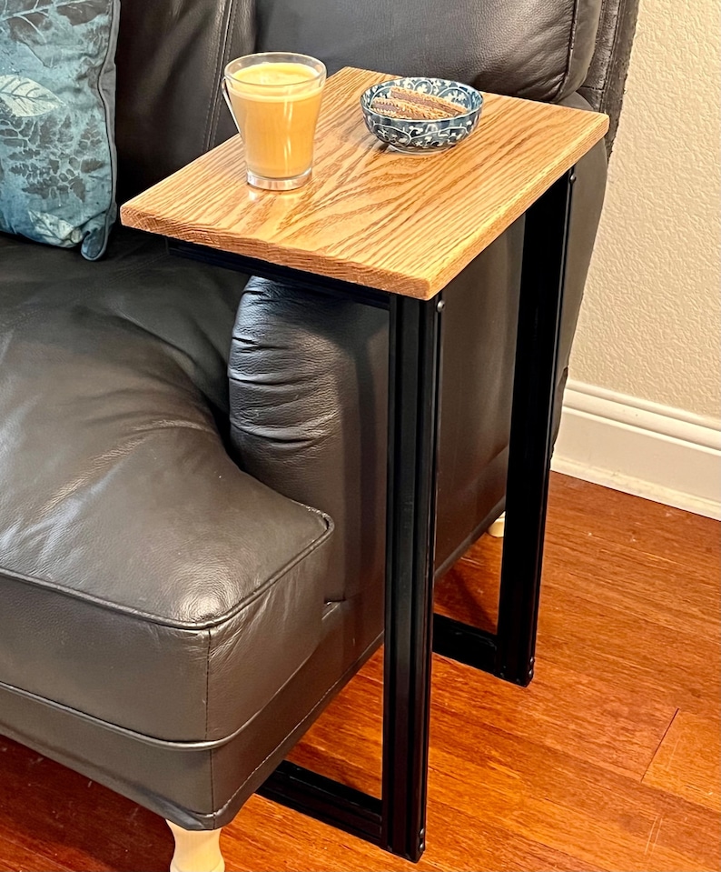 May include: A wooden side table with a light brown tabletop and black metal frame. The table is positioned next to a black leather couch. A glass of coffee and a small bowl are on the table.
