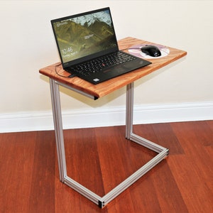 May include: A laptop computer sits on a wooden table with a silver metal frame. The table has a mouse pad and a wireless mouse. The time on the laptop screen is 10:46.