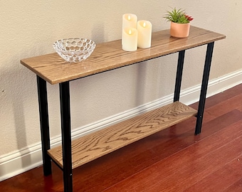 Puede incluir: Una mesa consola de madera con un marco de metal negro y un estante inferior. La mesa tiene un acabado de madera clara y está decorada con tres velas blancas y un cuenco de vidrio transparente.