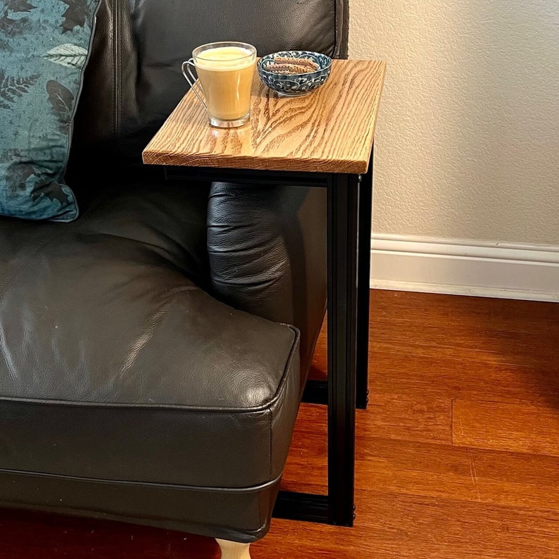 May include: A small, wooden side table with a light brown, rectangular top and black metal legs. A clear glass mug with a light-colored beverage and a small blue and white bowl are on the table. The table is next to a black leather couch.