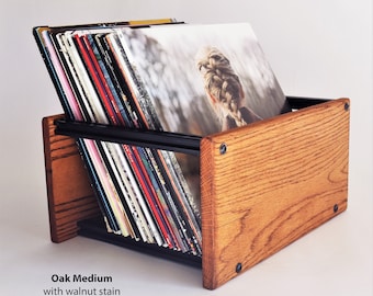 Puede incluir: Un soporte de discos de madera con tinte de nogal, que contiene varios discos de vinilo. El soporte está hecho de roble mediano y tiene soportes de metal negro. Los discos están en ángulo para facilitar la visualización y el acceso. El texto "Oak Medium with walnut stain" es visible.