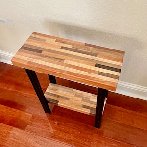 May include: A small wooden side table with a black metal frame. The tabletop and shelf are made of various wood types, creating a patchwork design.