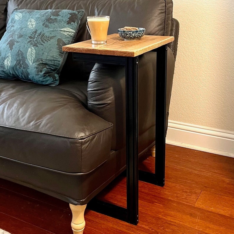 May include: A wooden side table with a black metal frame is positioned next to a brown leather sofa. The tabletop is made of light-colored wood. A cup of coffee and a small bowl are on the table. A teal pillow with a leaf pattern rests on the sofa.
