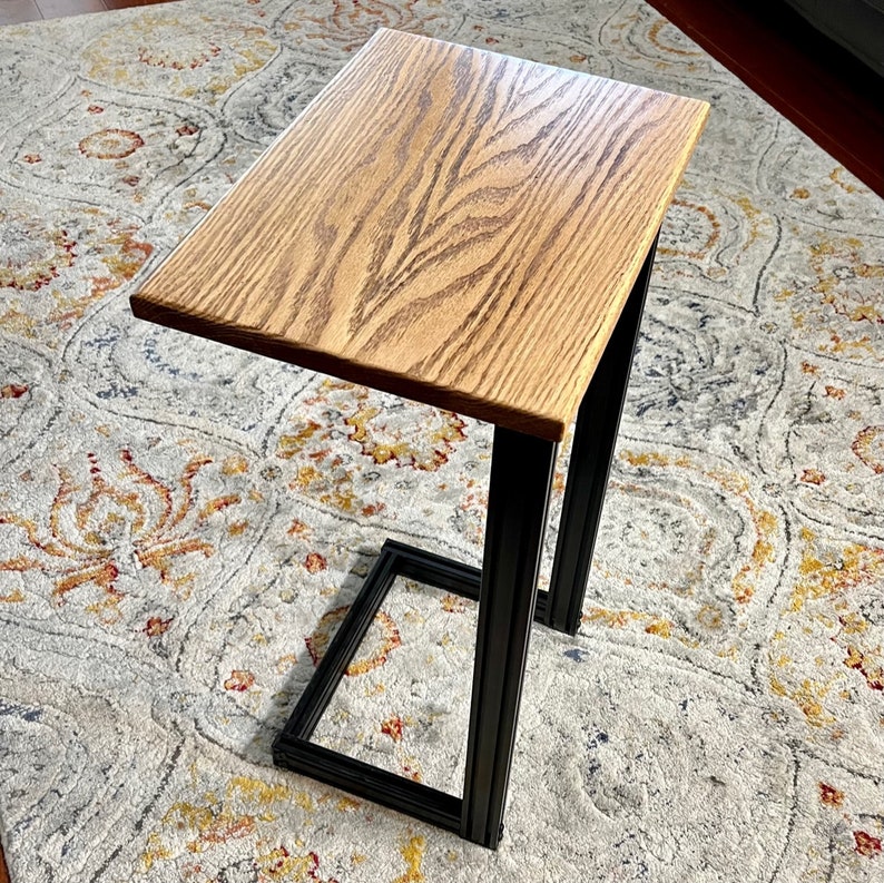 May include: A wooden side table with a light brown tabletop and black metal frame. The tabletop is rectangular and made of wood with a visible grain pattern. The frame is a black, geometric design. The table is designed to slide over a sofa or chair.