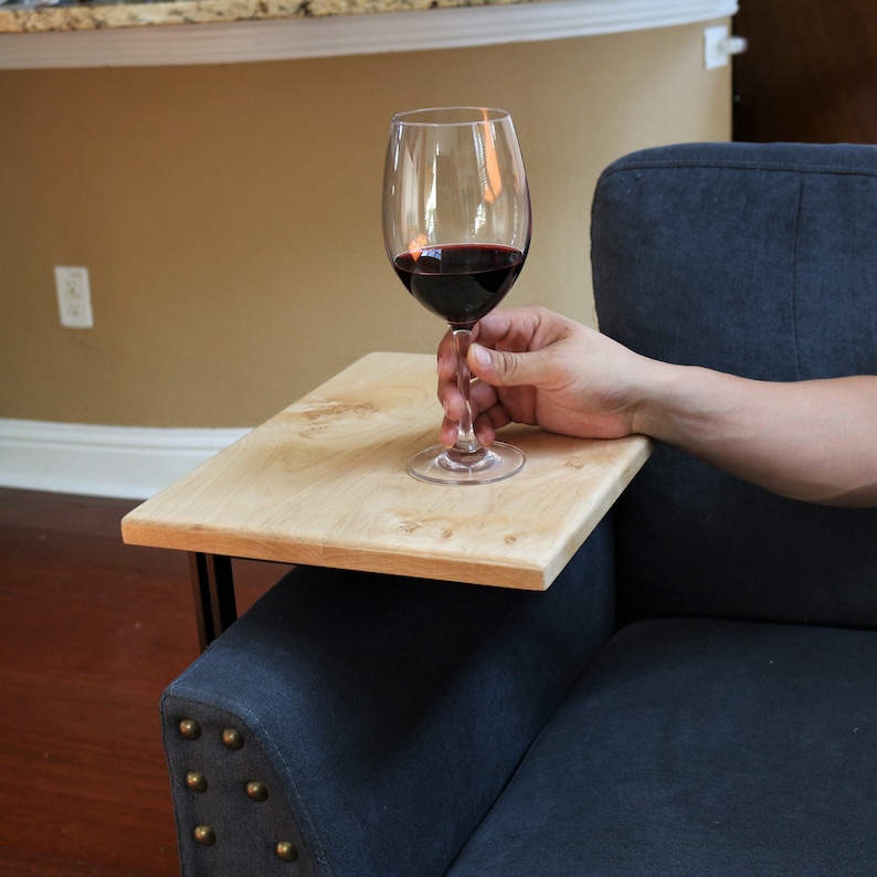 May include: A light-colored wooden sofa arm table with black metal legs. A clear wine glass filled with red wine is held above the table. The table is placed on a dark blue sofa.