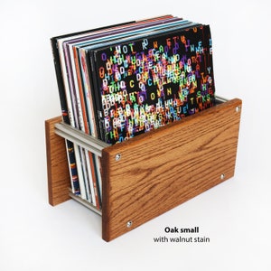 May include: A small oak record stand with a walnut stain finish. The stand is holding a stack of vinyl records with colorful, abstract designs.