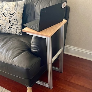 May include: A laptop sits on a wooden side table with a light brown top and silver metal frame. The table is positioned next to a black leather couch with a patterned pillow. The laptop is black.