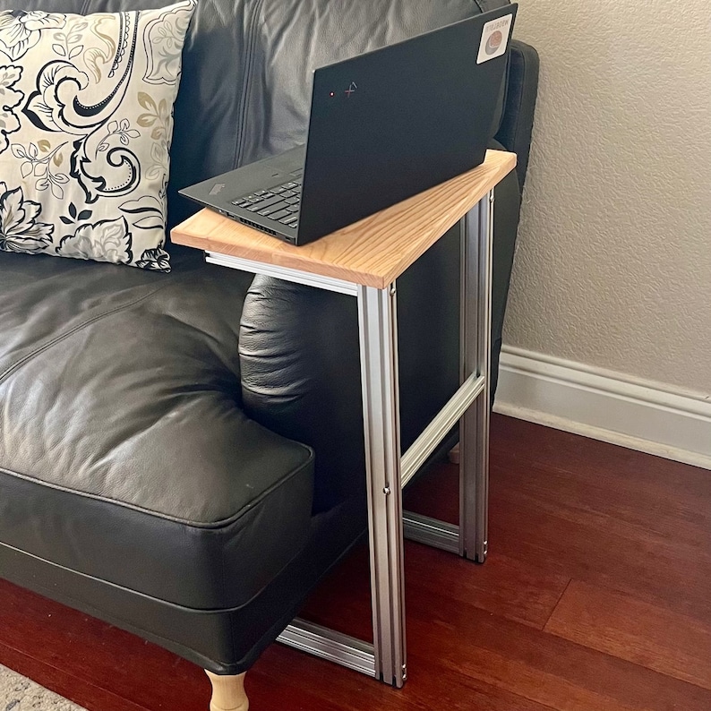 May include: A laptop sits on a wooden side table with a light brown top and silver metal frame. The table is positioned next to a black leather couch with a patterned pillow. The laptop is black.
