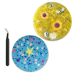 May include: Two round resin coasters with decorative elements. One coaster features sunflowers, bees, and yellow beads. The other has a blue background with a starfish, seashells, and letter beads. A black tweezer is on the left.
