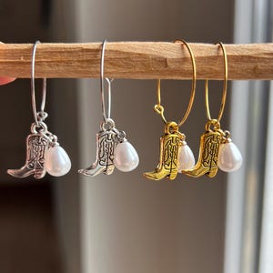 Può includere: Orecchini con un ciondolo a forma di stivale da cowboy e una perla bianca a forma di goccia. Disponibili in tonalità argento e oro, sono appesi a un cerchio. Gli orecchini sono esposti su un tassello di legno.