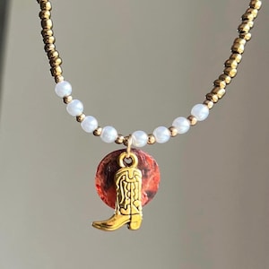 Peut inclure: Un collier en chaîne dorée avec des perles blanches et un pendentif rouge et or représentant une botte de cowboy.