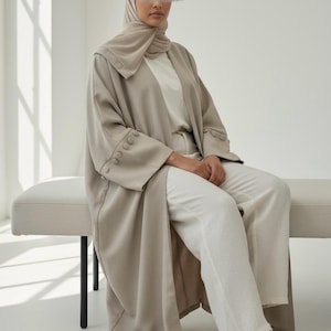 Peut inclure: Un abaya beige à devant ouvert avec un hijab assorti et un pantalon blanc. L'abaya comporte de gros boutons sur les manches. La personne porte des baskets blanches. L'ensemble est une tenue de mode modeste.