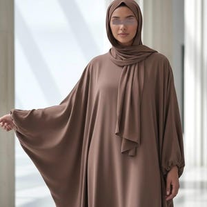 May include: A brown abaya dress with a matching hijab. The dress features wide sleeves and a flowing design. The model is wearing a head covering and a long, loose-fitting dress. The fabric appears to be a soft, draping material.