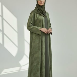 May include: An olive green abaya and matching hijab. The long-sleeved abaya has a textured pattern and is worn over a darker olive dress. The hijab is draped around the head and neck.