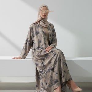 Puede incluir: Un vestido largo y fluido y una chaqueta a juego con la parte delantera abierta en una paleta de colores neutros con un estampado tie-dye. El conjunto se combina con un velo a juego y tacones altos color nude. El conjunto es adecuado para la moda modesta.