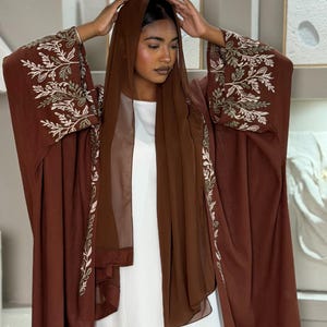 Puede incluir: Una abaya de color marrón oscuro abierta por delante con mangas largas y anchas que presentan un intrincado bordado floral en blanco y verde salvia en los puños y bordes frontales, junto a un pañuelo de gasa marrón y una prenda interior blanca.