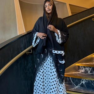 May include: A black abaya featuring a white polka dot pattern on the skirt and sleeve cuffs, accented with intricate black lace trim along the front opening. The garment includes a matching black head covering draped over the head and shoulders.