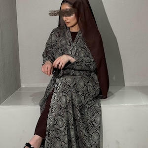Könnte beinhalten: Eine Frau trägt eine dunkelbraune Kopfbedeckung und einen langen, offen geschnittenen, gemusterten Robe. Die Robe hat ein geometrisches Design in Grau- und Schwarztönen. Das Outfit umfasst ein bordeauxfarbenes Oberteil und schwarze Sandalen.