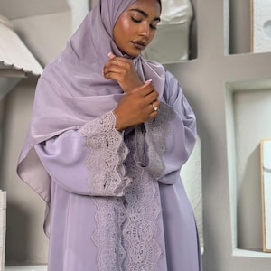 Puede incluir: Una abaya de color morado claro con mangas largas que presentan un borde de encaje ancho e intrincado en los puños y a lo largo de la abertura frontal. La prenda se combina con un hiyab morado claro a juego que cubre la cabeza y los hombros.