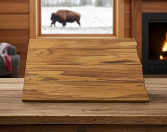 Può includere: Un tagliere in legno dalla forma unica, probabilmente un contorno statale, è appoggiato su un tavolo di legno rustico. La tavola presenta un motivo a venature naturali del legno ed è di colore marrone caldo. Lo sfondo include una finestra con un bisonte e un camino.