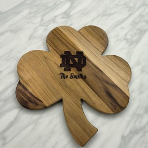 Pu&ograve; includere: Un tagliere in legno a forma di trifoglio con una venatura del legno marrone naturale. Il centro &egrave; inciso al laser con il logo del monogramma di Notre Dame e il testo The Smiths in un carattere marrone scuro, creando un netto contrasto.