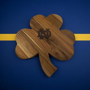 Pu&ograve; includere: Un tagliere in legno a forma di trifoglio caratterizzato da una venatura del legno marrone naturale e da un logo con monogramma di Notre Dame inciso in marrone scuro al centro. Il tagliere ha una finitura liscia e una sagoma a tre foglie.