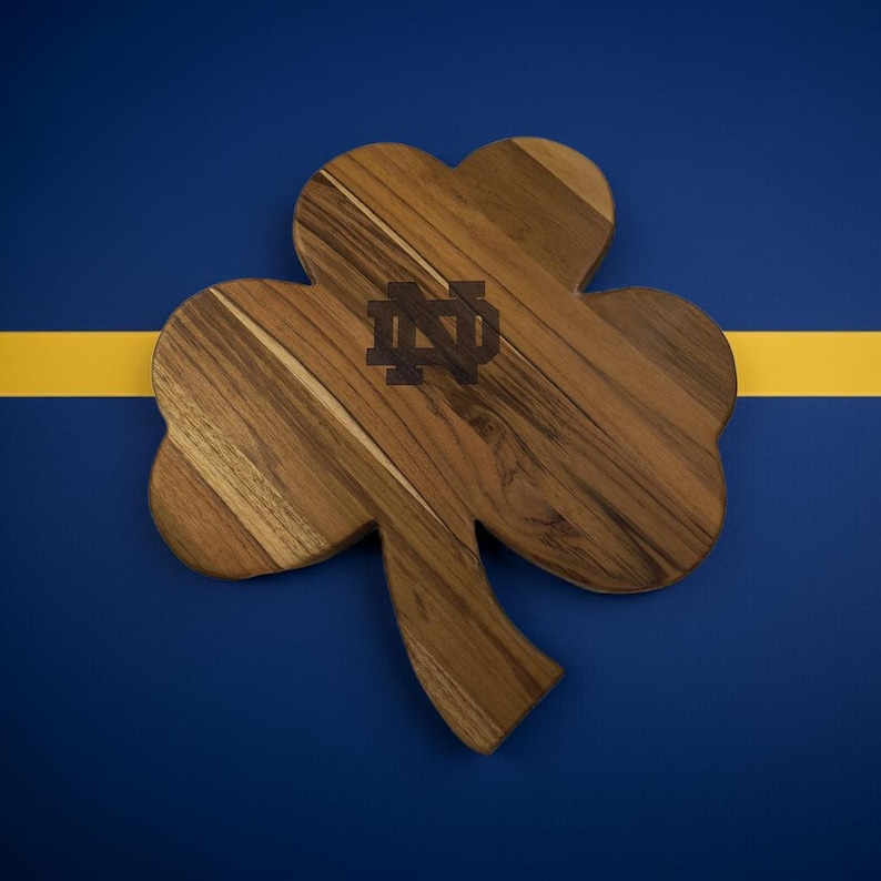 Pu&ograve; includere: Un tagliere in legno a forma di trifoglio caratterizzato da una venatura del legno marrone naturale e da un logo con monogramma di Notre Dame inciso in marrone scuro al centro. Il tagliere ha una finitura liscia e una sagoma a tre foglie.