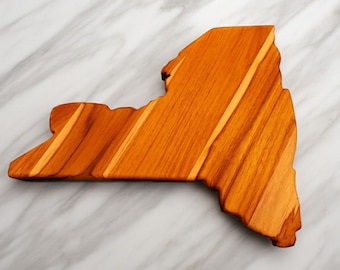 Può includere: Un tagliere in legno a forma dello stato di New York, caratterizzato da una venatura del legno naturale color ambra caldo con motivi lineari visibili e bordi lisci e rifiniti che seguono il distinto profilo geografico dello stato.