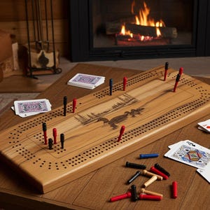 May include: A rectangular wooden cribbage board featuring a laser-engraved nature scene of ducks on a lake. The board has rows of drilled holes for scoring and includes several red, black, blue, and natural wood pegs along with playing cards.