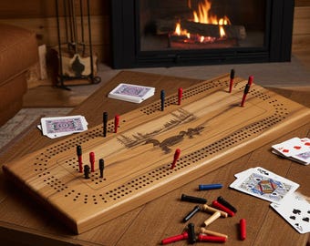 Può includere: Una tavola da cribbage rettangolare in legno con un'incisione laser raffigurante una scena naturale con anatre su un lago. La tavola presenta file di fori per il punteggio e include pioli rossi, neri, blu e in legno naturale e carte.