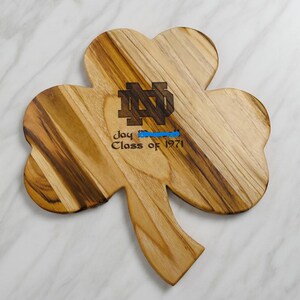 Pu&ograve; includere: Un tagliere in legno a forma di trifoglio con venature naturali. Al centro &egrave; inciso un logo monogramma ND scuro, un nome e la scritta Class of 1971. Il tagliere presenta una finitura liscia e un manico a forma di stelo.