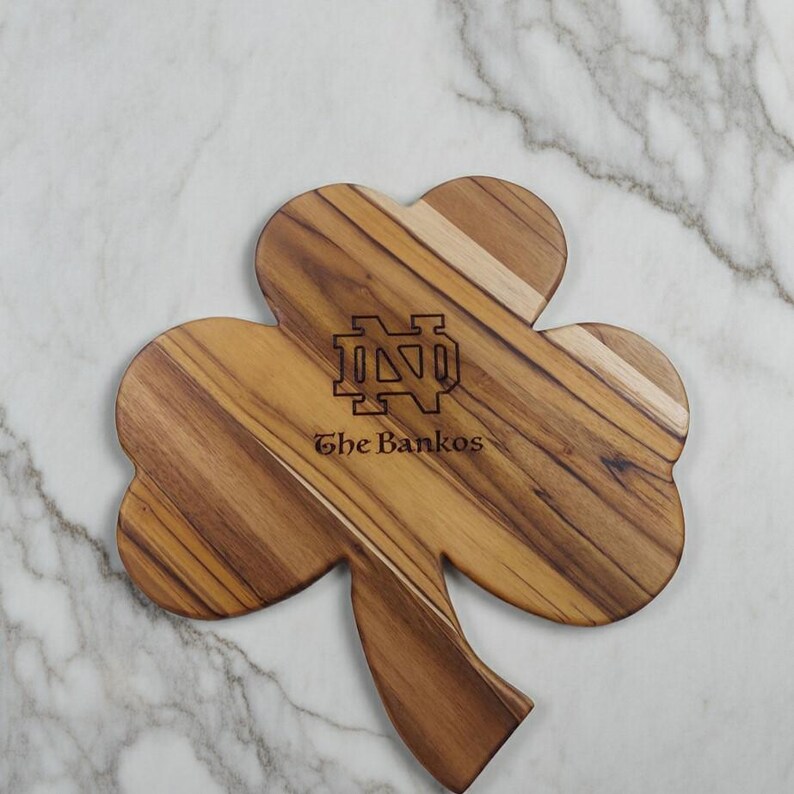 Pu&ograve; includere: Un tagliere in legno a forma di trifoglio con una venatura del legno marrone naturale. Il centro &egrave; inciso al laser con il logo del monogramma ND di Notre Dame sopra il testo The Bankos in un carattere serif scuro e in grassetto.