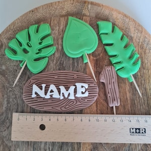 Könnte beinhalten: Ein Set aus fünf Tortendekorationen mit einem Holzschild mit dem Wort "NAME" in weißen Buchstaben, einer Nummer eins und drei grünen Blättern. Die Blätter haben verschiedene Formen, darunter ein Monstera-Blatt und ein herzförmiges Blatt.