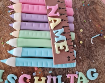 May include: A wooden cake board with a colorful cake decorated with fondant pencils, a ruler, and the words "NAME" and "SCHULTAG".