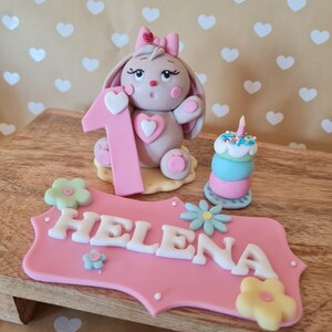 Op de afbeelding: Een roze taarttopper met een konijn dat een nummer één vasthoudt, een kleine taart met een kaars en een roze bord met de tekst "HELENA".