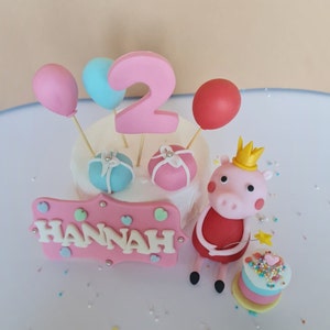 Könnte beinhalten: Eine rosa und weiße Geburtstagstorte, verziert mit einer rosa Schweinchenfigur, einer Zahl 2, Ballons und Geschenken. Die Torte ist außerdem mit einem rosa Schild verziert, auf dem "HANNAH" steht.