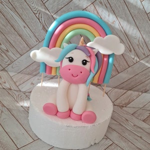 Könnte beinhalten: Ein weißer Einhorn-Kuchentopper mit einem rosa Gesicht, blauen und violetten Haaren und einem goldenen Horn. Das Einhorn sitzt vor einem Regenbogen mit weißen Wolken.