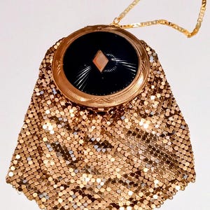 May include: A gold-tone metal mesh evening bag featuring a circular top frame with a black enamel inlay. The center of the black inlay displays a small diamond-shaped accent. The bag includes a thin gold-tone chain strap for carrying.