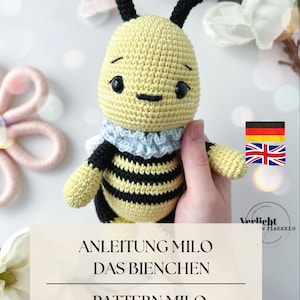 Könnte beinhalten: Ein gehäkeltes Amigurumi-Bienenspielzeug mit schwarzen und gelben Streifen, einem blau-weißen Rüschenkragen und schwarzen Augen. Das Spielzeug wird von einer Hand gehalten. Der Text "Anleitung Milo Das Bienchen" und "Pattern Milo The Bee" ist unter dem Spielzeug zu sehen.