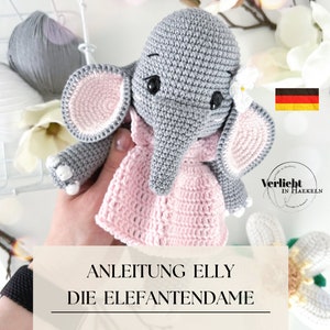 May include: A crocheted gray elephant wearing a pink dress with white accents. The elephant has black eyes and a white flower on its head. The text "Anleitung Elly Die Elefantendame" is visible in the image.
