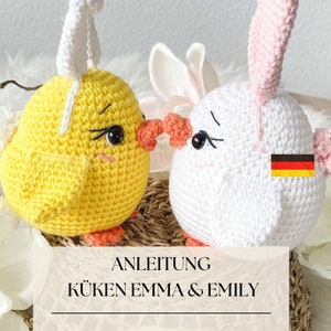 Puede incluir: Dos pollitos de crochet, uno amarillo y otro blanco, con orejas y picos rosas. Los pollitos están sentados en una cesta tejida y tienen un lazo blanco y rosa en la cabeza. El texto "ANLEITUNG KÜKEN EMMA & EMILY" está en un rectángulo blanco delante de los pollitos.